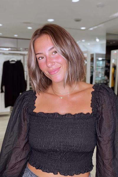 Eine Frau mit einem kinnlangen, aschblonden Balayage-Bob und dezentem Mittelscheitel steht in einem hellen Raum.