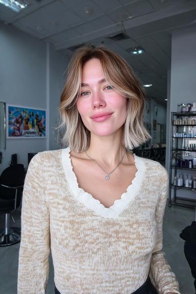 Eine Frau mit schulterlangem, aschblondem Balayage und weichen Wellen trägt einen Mittelscheitel.