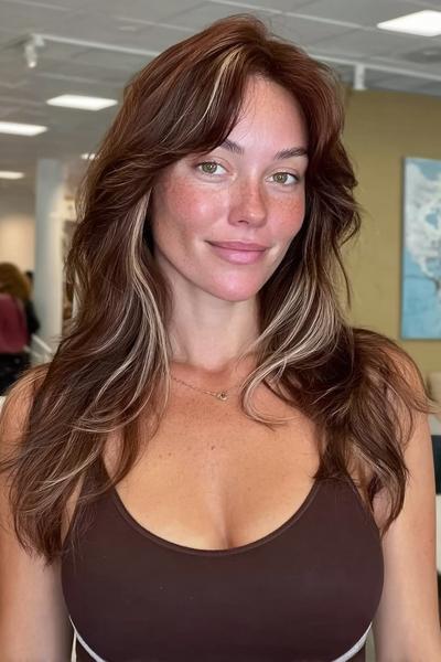 Eine Frau mit langen, braunen Haaren und blonden Highlights trägt eine sanfte, wellige Stufenschnitt-Frisur mit Mittelscheitel.