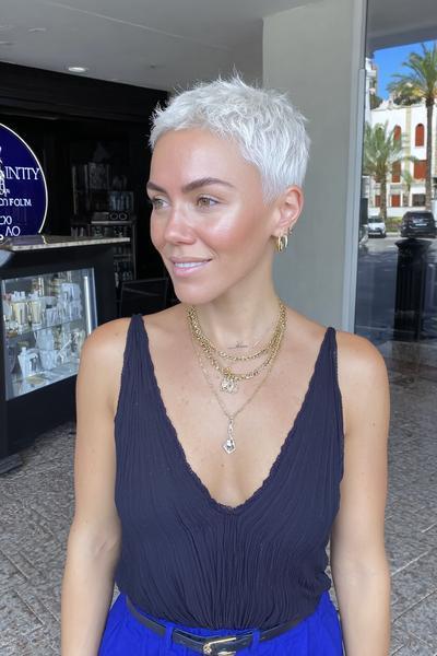 Eine Frau mit einem sehr kurzen, platinblonden Pixie-Haarschnitt und seitlich gelegtem Pony steht draußen vor einem Gebäude.