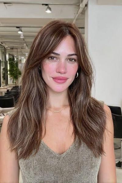 Eine Frau mit langen, braun-blonden Haaren im Balayage-Stil und sanften Wellen trägt einen Mittelscheitel.