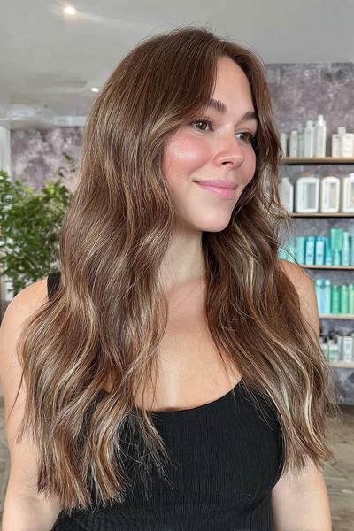 Eine Frau mit langen, welligen Haaren in einem Braunton mit sanften blonden Balayage-Highlights.
