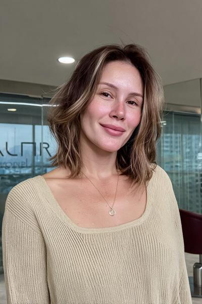 Eine Frau mit schulterlangem, dunkelblondem Haar trägt eine natürliche, leicht gewellte Frisur mit sanften Highlights.