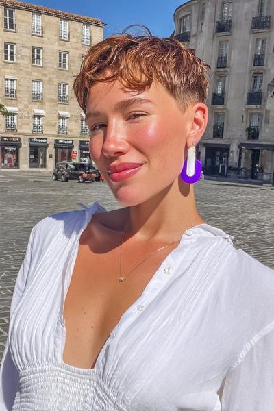 Eine Frau mit einem kurzen, welligen Pixie Cut und seitlich gelegtem Pony steht im Sonnenschein auf einem Stadtplatz.