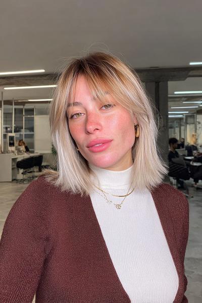 Eine Frau mit einem kinnlangen, glatten Balayage-Haarschnitt in Aschblond und fransigem Pony steht in einem modernen Friseursalon.