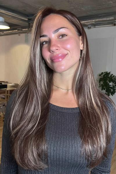 Eine Frau mit langen, glatten Haaren in einem Braunton mit dezenten, blonden Highlights trägt einen Mittelscheitel.