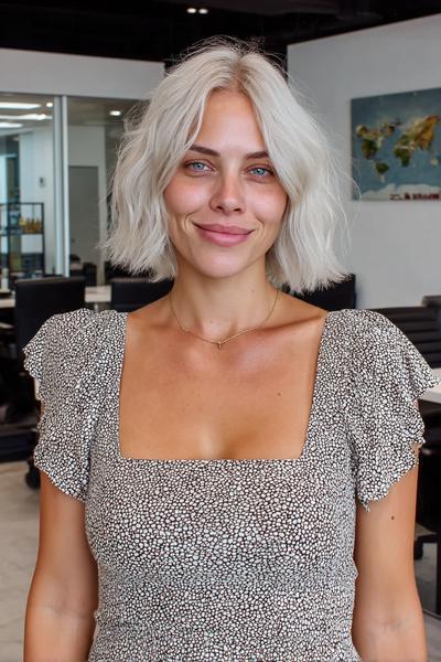 Eine Frau mit welligem, kinnlangem aschblondem Balayage-Haar trägt ein gemustertes Kleid und steht in einem modernen Büro.