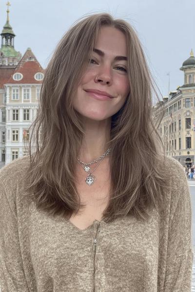 Eine Frau mit mittel-langem, dunkelblondem Haar, das in sanften Stufen und mit einem leichten Mittelscheitel locker und natürlich fällt.