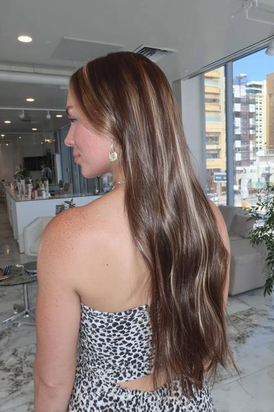 Frau mit langen, glatten braunen Haaren und dezenten, hellblonden Balayage-Highlights steht in einem modernen Friseursalon.