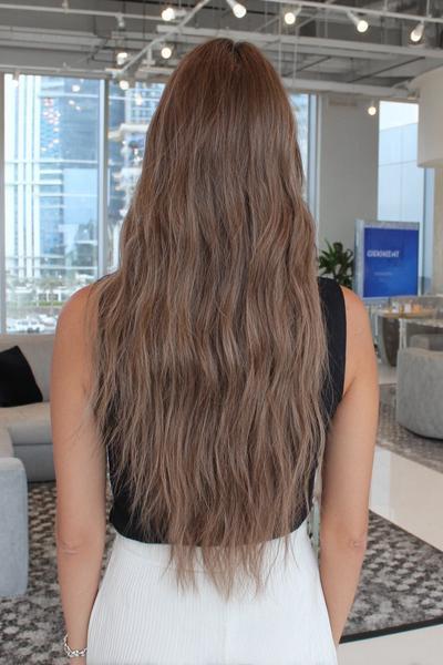 Eine Frau mit langen, welligen haaren in einem natürlichen Braun-Blond-Ton steht mit dem Rücken zur Kamera in einem modernen Innenraum.
