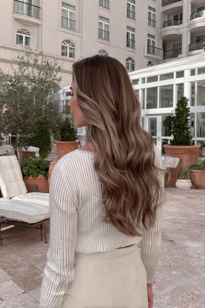 Eine Frau mit langen, lockigen Haaren in einem natürlichen Braun-Blond-Verlauf (Balayage) steht im Freien und trägt einen weißen Strickpullover.