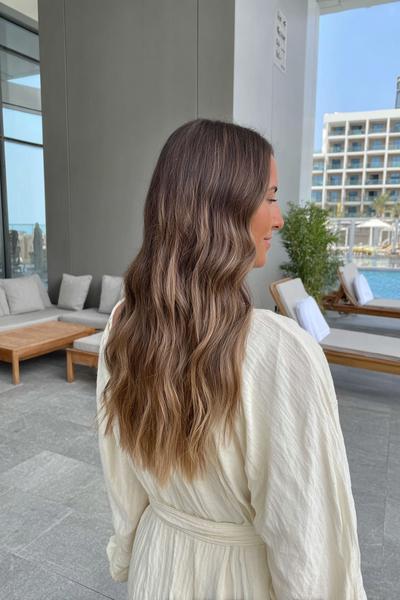 Eine Frau mit langen, lockeren Haaren im Braun-Blond-Balayage-Stil steht in einem modernen Außenbereich.