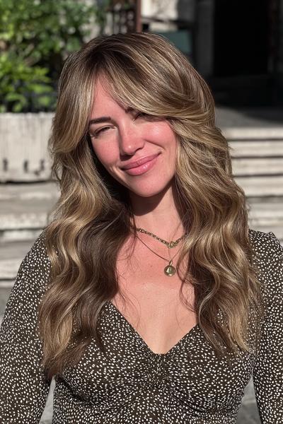 Eine Frau mit langen, welligen, dunkelblonden Haaren und sanften Balayage-Highlights steht im Sonnenlicht.