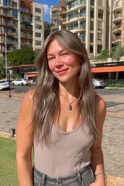 Eine Frau trägt lange, leicht gewellte Haare in einem aschblonden Balayage-Stil mit dunklem Ansatz und natürlichen Highlights.