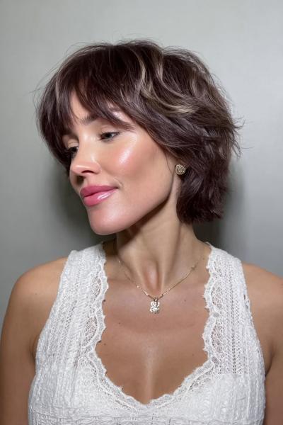 Eine Frau mit einem kurzen, modernen Pixie-Cut mit leichtem Undercut und seitlichem Pony sitzt lächelnd im Raum.