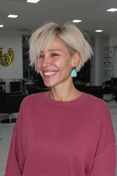 Eine lächelnde Frau mit einem blonden, welligen Pixie Cut mit langen, seitlichen Ponysträhnen steht in einem modernen Friseursalon.