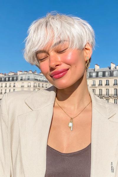 Eine Frau mit einem modernen, weißen, kurzen Pixie-Cut mit fransigem Pony steht lächelnd im Sonnenlicht.