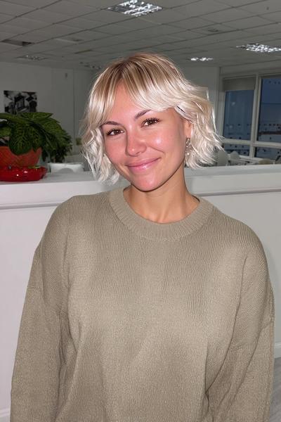 Eine Frau mit einem welligen, kinnlangen, blonden Pixie Cut mit Pony steht in einem modernen Innenraum.