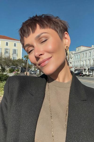 Eine Frau mit einem kurzen, dunkelbraunen Pixie Cut und fransigem Pony posiert draußen bei sonnigem Wetter.
