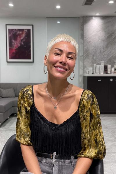 Eine Frau mit einem sehr kurzen, platinblonden Pixie-Cut und seitlich getrimmten Konturen sitzt lächelnd in einem modernen Salon.