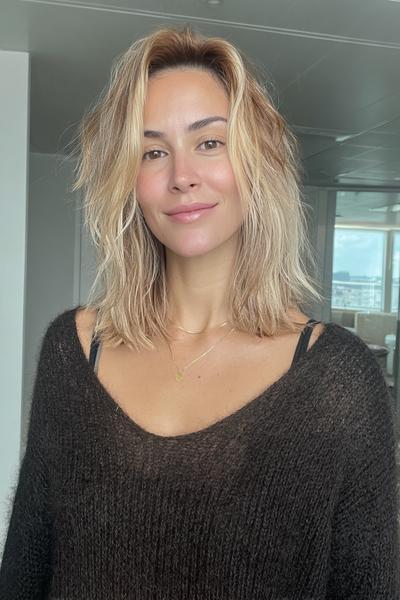 Eine Frau mit schulterlangem, aschblondem Balayage-Haar und leicht gewellter Struktur steht in einem hellen Raum.