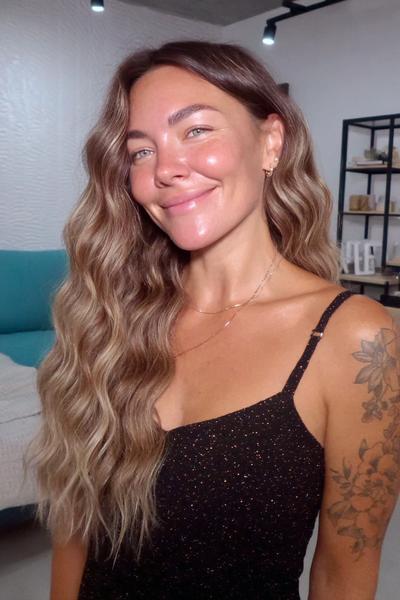 Eine Frau mit langen, gewellten, dunkelblonden Haaren trägt ein schwarzes Top und lächelt in einem modern eingerichteten Raum.
