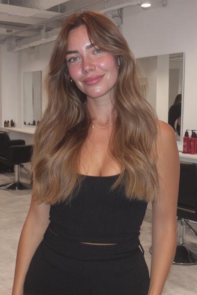 Eine Frau mit langen, welligen dunkelblonden Haaren und weich fallendem Mittelscheitel steht in einem modernen Friseursalon.
