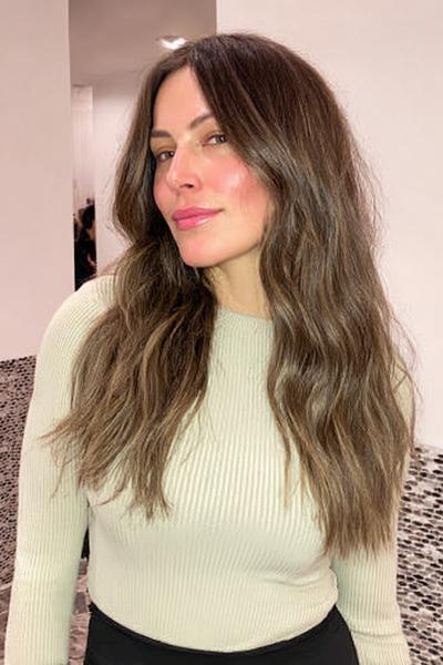 Eine Frau mit langen, gewellten Haaren in einem natürlichen Braun-Blond-Ton und Mittelscheitel.