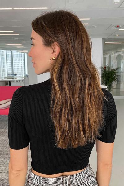 Eine Frau mit langen, welligen braun-blonden Haaren und dezenten Balayage-Strähnen trägt ein schwarzes, geripptes Oberteil.