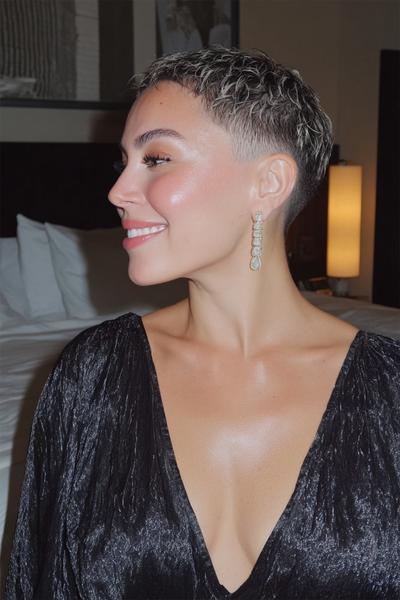 Eine Frau mit einem kurz geschnittenen, platinblonden Pixie Cut mit seitlichem Pony sitzt draußen und lächelt.