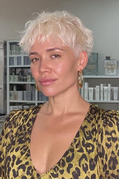 Eine Frau mit einem modernen, blond platinfarbenen Pixie Cut mit strukturierten, leicht gewellten und fransigen Spitzen steht in einem Friseursalon.