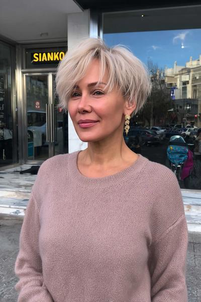 Eine Frau mit einem modernen, blonden Pixie-Cut mit fransigem Pony und längeren Strähnen an den Seiten steht vor einem Laden.