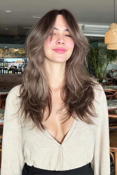 Eine Frau mit langen, dunkelblonden Haaren trägt eine moderne Stufenschnitt-Frisur mit sanften Wellen und Mittelscheitel.