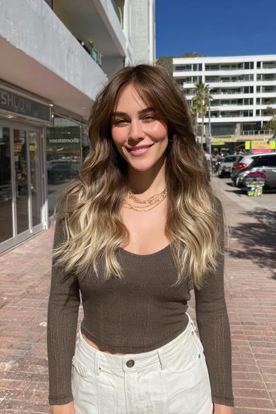 Eine Frau mit langen, welligen Haaren trägt ein Balayage in Aschblond, wobei die Haarspitzen deutlich heller als der dunklere Ansatz sind.